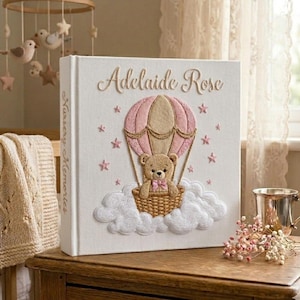 Puede incluir: Un &aacute;lbum de recuerdos para beb&eacute;s blanco con el nombre "Adelaide Rose". La portada presenta un osito de peluche en un globo aerost&aacute;tico con detalles en rosa y estrellas. El lomo dice "Nursery Memories".