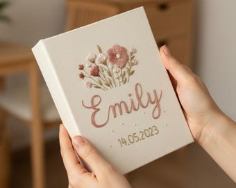 May include: A cream-colored, rectangular box with embroidered floral details and the name "Emily" in a cursive font. The embroidery includes pink and red flowers, with the date "14.05.2023" below the name.