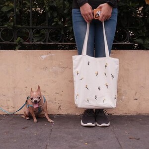 May include: A white tote bag with a floral pattern, a small brown dog on a leash, and a person wearing blue jeans and black shoes.