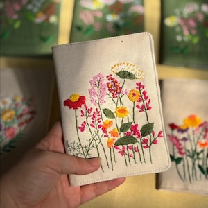 Könnte beinhalten: Ein beiger Reisepass-Umschlag mit einem gestickten Blumendesign mit roten, rosa, gelben und orangen Blumen mit grünen Stielen und Blättern. Weitere Umschläge mit ähnlichen Designs sind im Hintergrund zu sehen.