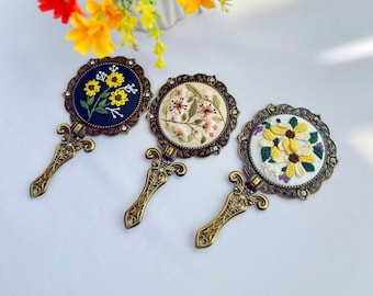 Könnte beinhalten: Drei Handspiegel im antiken Stil mit gestickten Blumenmustern. Jeder Spiegel hat einen bronzefarbenen Rahmen und Griff. Die gestickten Designs umfassen Sonnenblumen und andere Blumen in Gelb, Rosa und Weiß.