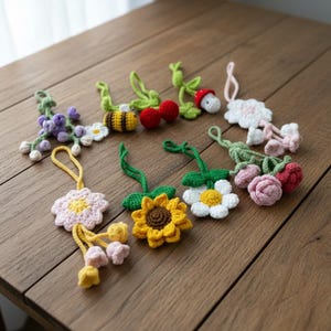 Op de afbeelding: Een verzameling handgemaakte gehaakte bloemen- en fruitornamenten in verschillende kleuren. De ornamenten omvatten een gele zonnebloem, een witte madelief, een roze roos, rode kersen en een bij. Ze zijn gerangschikt op een houten oppervlak.