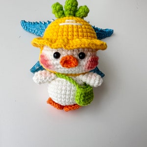 May include: A crocheted stuffed toy chick wearing a yellow hat with a green top and a green and white bag. The chick has a white body, orange beak, and black eyes.
