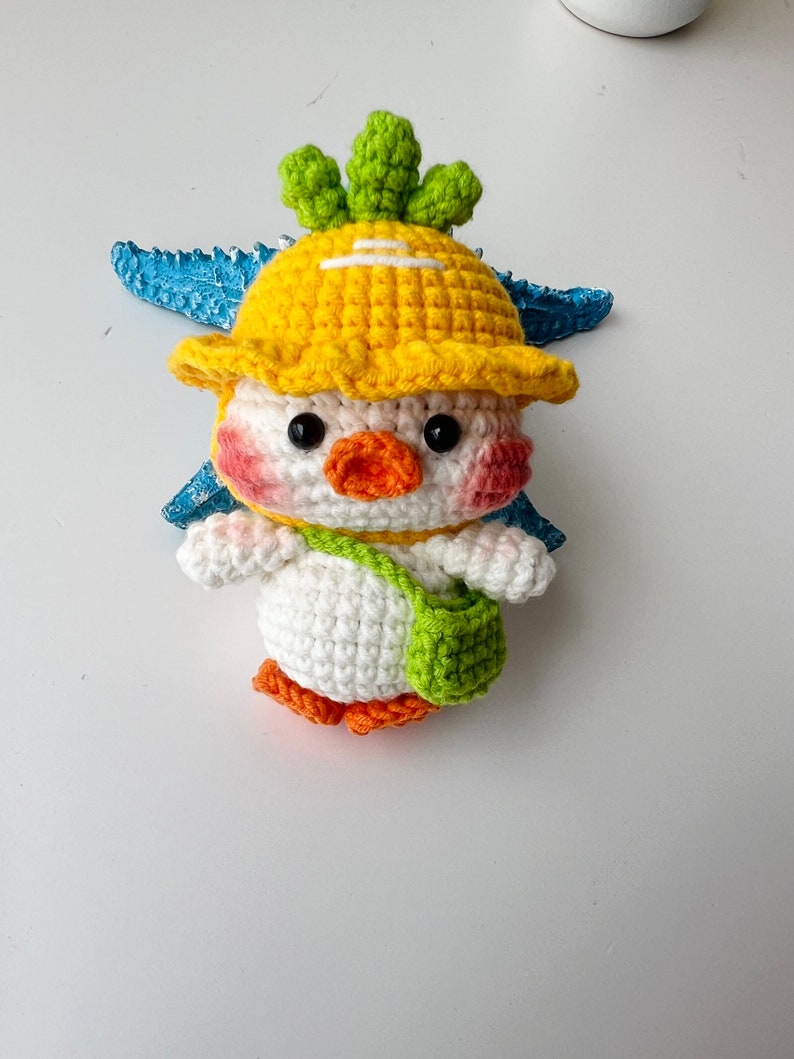 May include: A crocheted stuffed toy chick wearing a yellow hat with a green top and a green and white bag. The chick has a white body, orange beak, and black eyes.