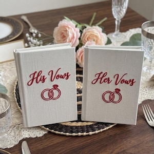Pode incluir: Dois livros revestidos de tecido na cor off-white posicionados verticalmente. O livro da esquerda apresenta o texto bordado em vermelho His Vows acima de um &iacute;cone de dois an&eacute;is bordados em vermelho. O livro da direita exibe o texto Her Vows.