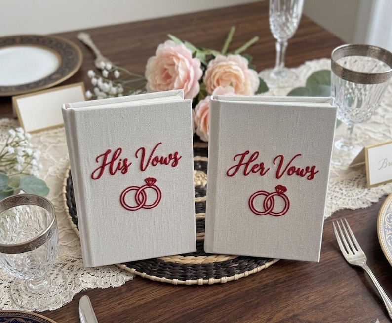 Pode incluir: Dois livros revestidos de tecido na cor off-white posicionados verticalmente. O livro da esquerda apresenta o texto bordado em vermelho His Vows acima de um &iacute;cone de dois an&eacute;is bordados em vermelho. O livro da direita exibe o texto Her Vows.