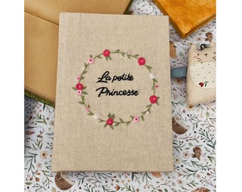 May include: A beige linen notebook with a floral embroidery design. The embroidery is a wreath of pink flowers with green leaves and the text "La petite Princesse" in black thread.