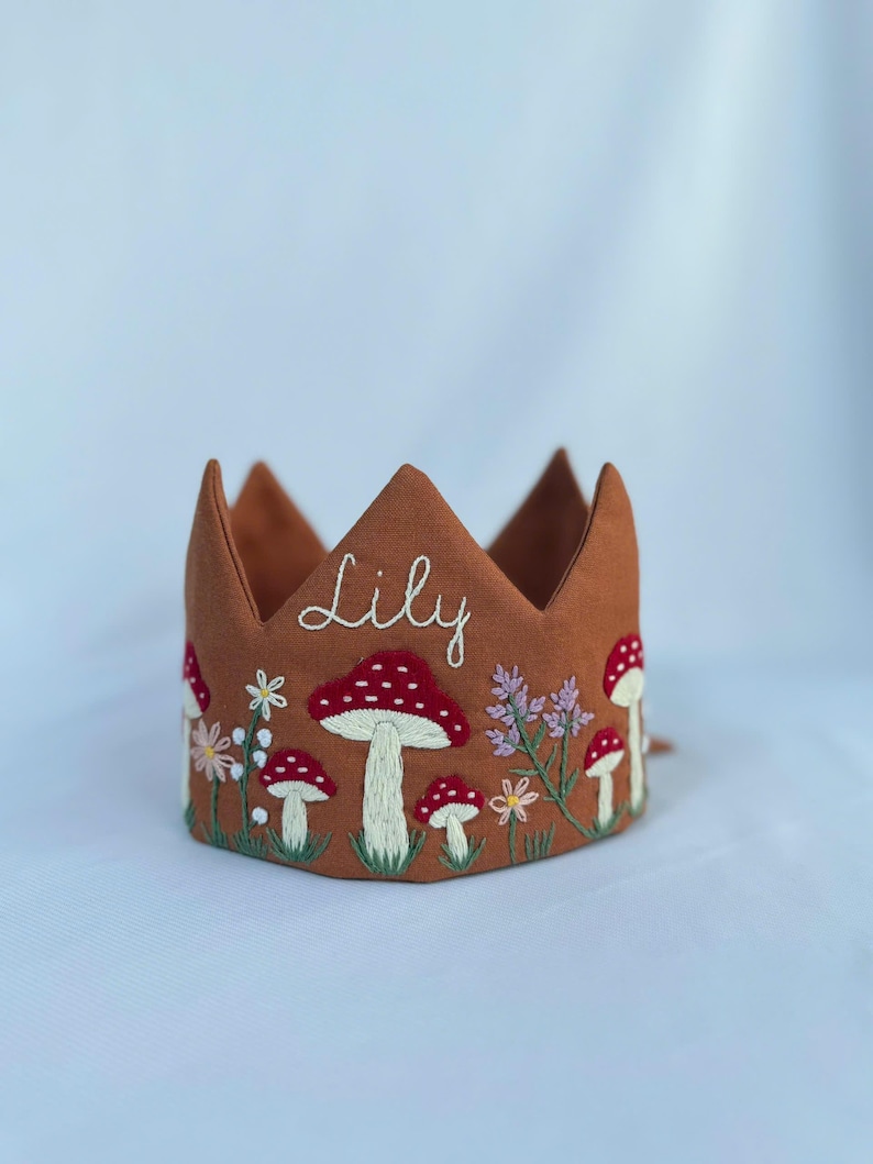 May include: A brown fabric crown embroidered with red and white mushrooms, flowers, and green grass. The name "Lily" is embroidered in white script across the front. The crown has pointed peaks.