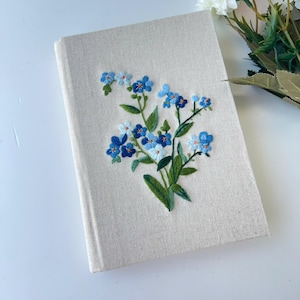 Könnte beinhalten: Ein beigefarbenes Leinen-Tagebuch mit blauen und weißen gestickten Vergissmeinnicht-Blumen.
