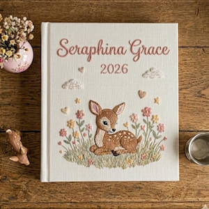 Puede incluir: Diario bordado con el nombre "Seraphina Grace" y el a&ntilde;o 2026 en hilo rosa. La portada presenta un cervatillo rodeado de flores y nubes. El diario est&aacute; sobre una superficie de madera.