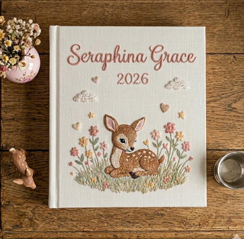 Puede incluir: Diario bordado con el nombre "Seraphina Grace" y el a&ntilde;o 2026 en hilo rosa. La portada presenta un cervatillo rodeado de flores y nubes. El diario est&aacute; sobre una superficie de madera.