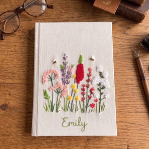 Op de afbeelding: Een crèmekleurig notitieboek met een geborduurd bloemmotief met roze, paarse, rode en gele bloemen, met twee bijen. De naam "Emily" is in groen schrift eronder geborduurd. Het notitieboek ligt op een houten oppervlak.
