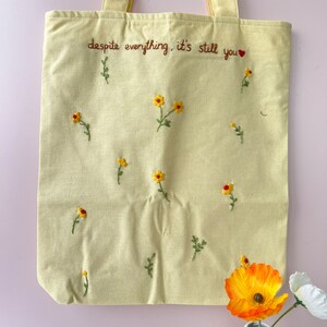 May include: A yellow canvas tote bag with the embroidered text "despite everything, it's still you" and small yellow flowers.