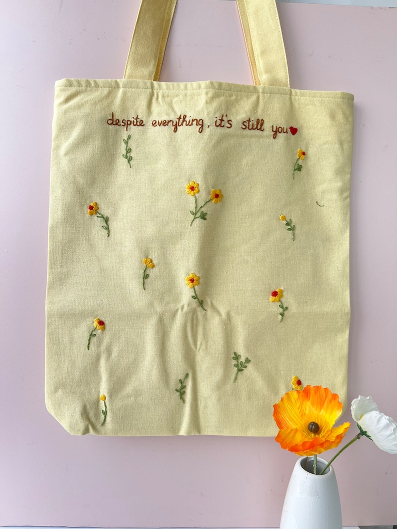 May include: A yellow canvas tote bag with the embroidered text "despite everything, it's still you" and small yellow flowers.