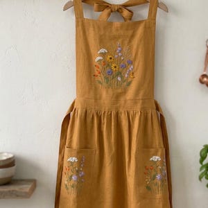 May include: A mustard yellow apron featuring intricate floral embroidery on the chest and two front pockets. The design includes colorful wildflowers in shades of white, orange, purple, and yellow. The apron has wide shoulder straps and a tie back.