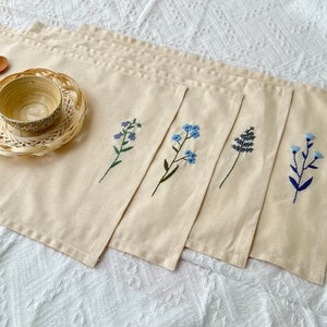 Könnte beinhalten: Set aus fünf beigefarbenen Tischsets aus Leinen mit gestickten Blumenmotiven. Jedes Tischset zeigt ein anderes blaues Blumendesign.