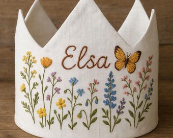May include: A white fabric crown with a floral embroidery design. The crown features the name "Elsa" in brown script, a butterfly, and colorful embroidered flowers. The crown has a round base and pointed top.