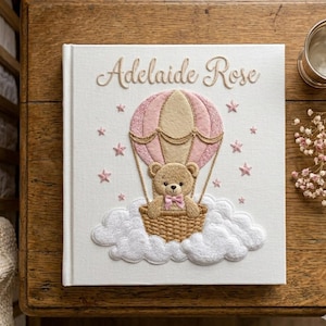 Puede incluir: Un libro de recuerdos para beb&eacute;s blanco con el nombre "Adelaide Rose" bordado en oro. La portada presenta un globo aerost&aacute;tico rosa con un osito de peluche en una cesta, rodeado de nubes y estrellas rosas. Un recuerdo encantador.