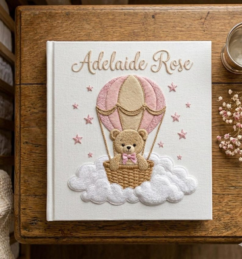 Puede incluir: Un libro de recuerdos para beb&eacute;s blanco con el nombre "Adelaide Rose" bordado en oro. La portada presenta un globo aerost&aacute;tico rosa con un osito de peluche en una cesta, rodeado de nubes y estrellas rosas. Un recuerdo encantador.