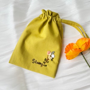 May include: A yellow fabric drawstring bag with a floral embroidery design and the name "Stacey" stitched on the front.