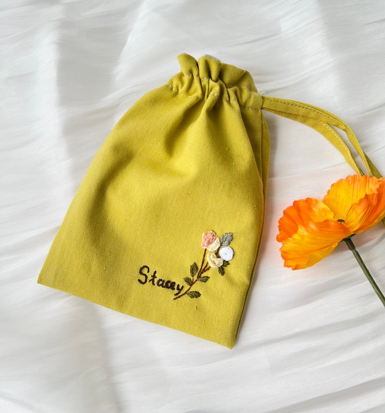 May include: A yellow fabric drawstring bag with a floral embroidery design and the name "Stacey" stitched on the front.