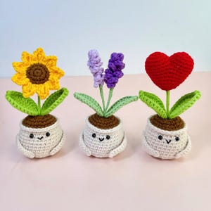 May include: Three crocheted flower pots with smiling faces. One pot has a yellow sunflower, another has purple and lavender flowers, and the third has a red heart-shaped flower. Each pot is beige with brown "soil" and green leaves.