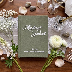 Pode incluir: Um livro de visitas revestido em tecido verde-oliva com texto bordado em branco que diz Michael + Sarah, com a data 27/06/26 e a localiza&ccedil;&atilde;o Mount Desert, Maine, bordados em fio branco na parte inferior da capa frontal.