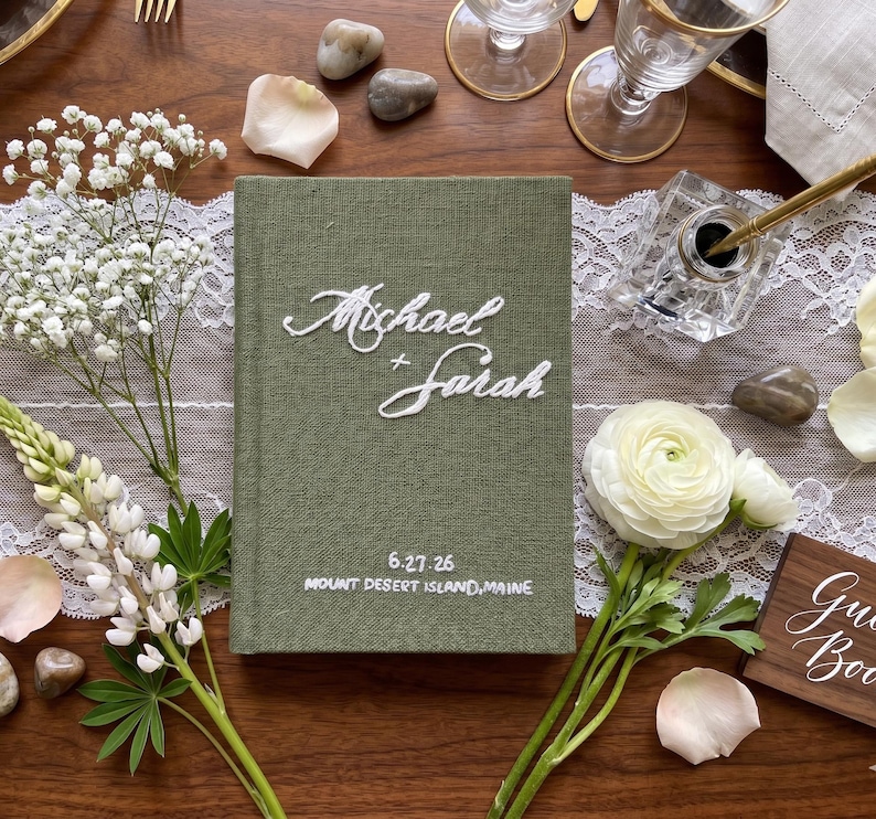 Pode incluir: Um livro de visitas revestido em tecido verde-oliva com texto bordado em branco que diz Michael + Sarah, com a data 27/06/26 e a localiza&ccedil;&atilde;o Mount Desert, Maine, bordados em fio branco na parte inferior da capa frontal.
