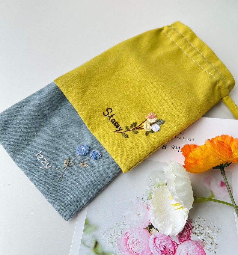May include: Two small fabric pouches with embroidered flowers and names. The pouch on the left is blue with the name "Izzy" and the pouch on the right is yellow with the name "Stacey".