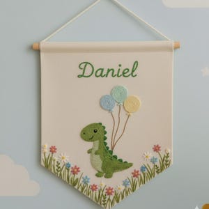 Op de afbeelding: Geborduurde banner in crème met de naam "Daniel" in groene letters. Een groene dinosaurus met drie pastelkleurige ballonnen is afgebeeld. De banner heeft een houten stok en een touwtje om op te hangen. Bloemenborduurwerk onderaan.