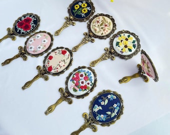 Könnte beinhalten: Eine Sammlung verzierter Handspiegel mit gestickten Blumenmustern. Jeder Spiegel zeigt ein anderes Blumenmuster, darunter Rosen, Sonnenblumen und Gänseblümchen, in einem ovalen oder runden Rahmen mit einem dekorativen Griff. Die Spiegel haben eine Vintage-Ästhetik.