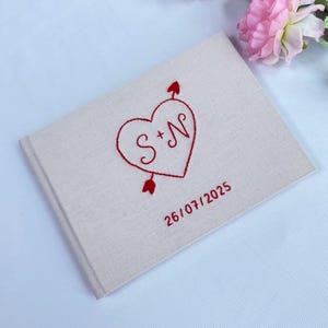 Peut inclure: Un livre d'or rectangulaire recouvert de tissu beige avec des détails brodés en rouge. La couverture présente un cœur traversé par une flèche avec les lettres S + N à l'intérieur, et la date 26/07/2025 brodée en fil rouge sous le cœur.
