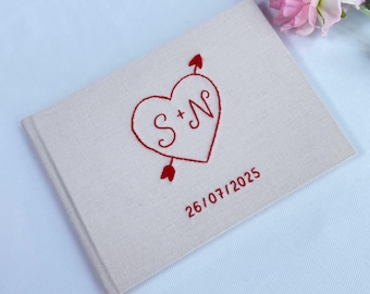 May include: A rectangular beige fabric-covered guest book featuring red embroidered details. The cover displays a heart pierced by an arrow with the letters S + N inside, and the date 26/07/2025 embroidered in red thread below the heart.
