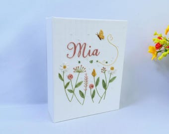 May include: A white rectangular fabric-covered box featuring embroidered floral designs in green, pink, yellow, and white. The name Mia is embroidered in a cursive pink font above the flowers, accompanied by a small yellow butterfly and swirls.