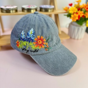 May include: A gray baseball cap with embroidered flowers in shades of blue, orange, and yellow. The phrase "stay wild" is embroidered in black script below the flowers. The cap is made of a soft, textured fabric.