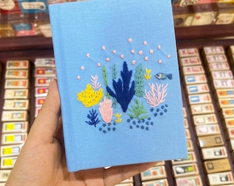May include: A light blue notebook with a hand-stitched design of a coral reef with pink, yellow, green, and blue coral, seaweed, and a small blue fish.