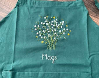 May include: A dark green apron with a pocket, embroidered with a bouquet of white flowers and yellow stars. The name "Mags" is embroidered below the flowers.