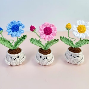 May include: Three crocheted flower pot dolls. Each doll has a different coloured flower: blue, pink, and white. The pots are white with a smiling face and brown soil. The flowers have green leaves and stems.