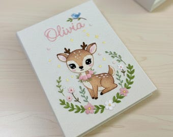May include: A light-colored book with the name "Olivia" in pink script. The cover features a cartoon deer with a floral garland, surrounded by a wreath of leaves and flowers. A blue bird sits above the name.