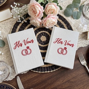 Pode incluir: Dois livros de votos de capa dura branca com capas texturizadas. O livro da esquerda apresenta o texto bordado em vermelho His Vows acima de um &iacute;cone de an&eacute;is vermelhos. O livro da direita apresenta o texto bordado em vermelho Her Vows acima de um &iacute;cone de an&eacute;is vermelhos.