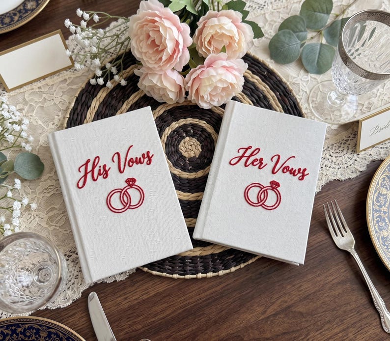 Pode incluir: Dois livros de votos de capa dura branca com capas texturizadas. O livro da esquerda apresenta o texto bordado em vermelho His Vows acima de um &iacute;cone de an&eacute;is vermelhos. O livro da direita apresenta o texto bordado em vermelho Her Vows acima de um &iacute;cone de an&eacute;is vermelhos.