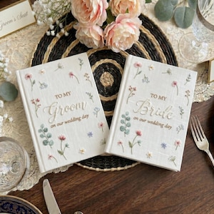 May include: Two white wedding guest books with floral embroidery. One book reads "TO MY Groom on our wedding day," and the other reads "TO MY Bride on our wedding day." The books are on a table with floral arrangements.