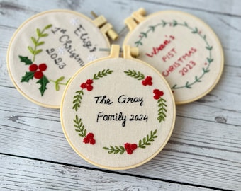 Può includere: Tre ornamenti natalizi ricamati con sfondo bianco e ricami verdi e rossi. Gli ornamenti sono circolari e hanno una cornice in legno. Il testo sugli ornamenti recita "Elige's 1st Christmas 2023", "Liam's First Christmas 2023" e "The Gray Family 2024".