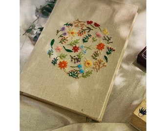 May include: A beige linen journal with a circular embroidery design of colorful flowers and leaves. The embroidery is in shades of red, orange, yellow, green, blue, and purple.