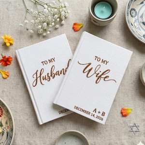 Pode incluir: Dois livros de capa dura branca com superf&iacute;cies texturizadas. Um livro apresenta o texto castanho TO MY Husband, e o outro apresenta TO MY Wife, A + B, e DECEMBER 14, 2026. Os livros est&atilde;o posicionados lado a lado numa superf&iacute;cie de tecido.
