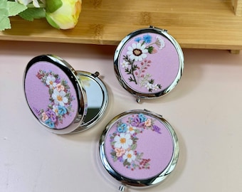 Könnte beinhalten: Drei runde Kompaktspiegel mit silbernen Rändern und gestickten Blumenmustern auf einem hellvioletten Hintergrund. Die Stickerei zeigt weiße, blaue und rosa Blumen. Ein Spiegel ist geöffnet und zeigt die reflektierende Oberfläche.