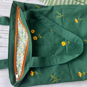May include: A dark green tote bag with a zipper closure and embroidered yellow flowers. The bag has a brown zipper pull and a floral patterned lining.
