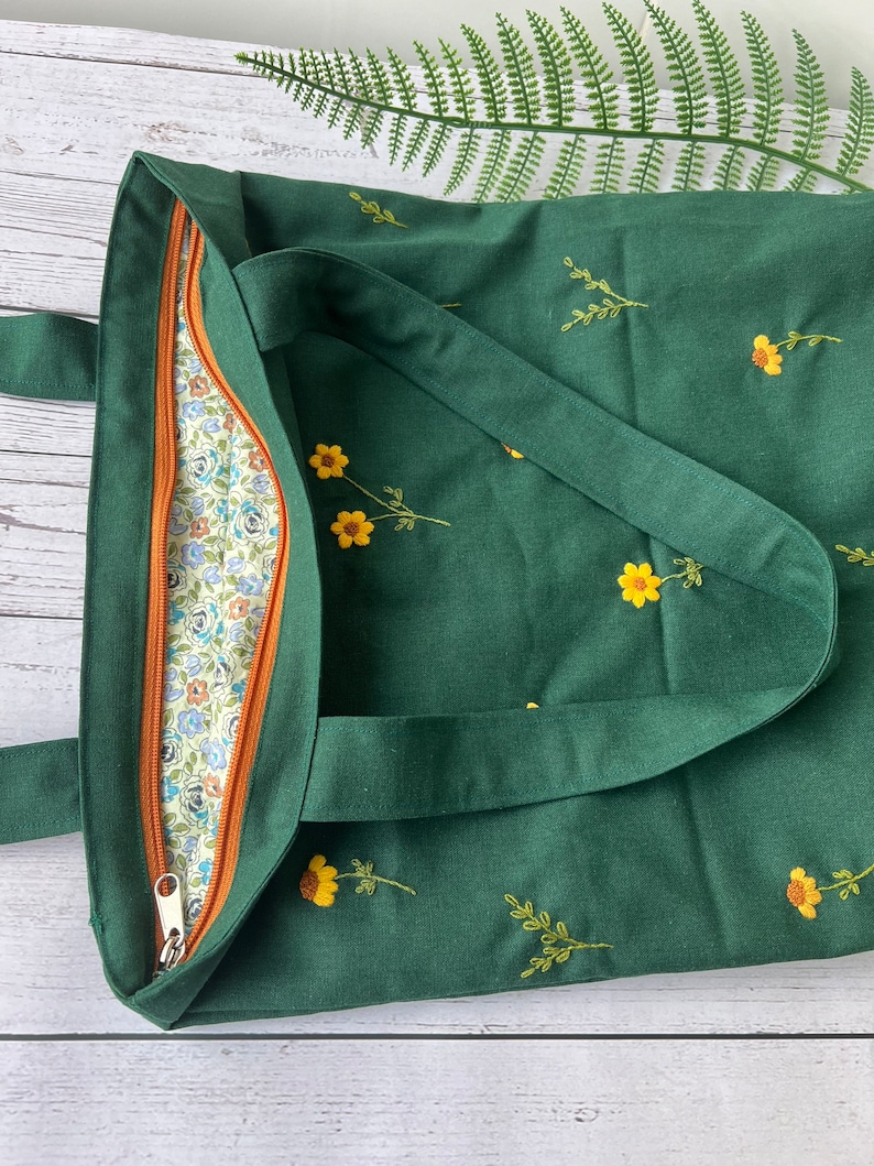 May include: A dark green tote bag with a zipper closure and embroidered yellow flowers. The bag has a brown zipper pull and a floral patterned lining.
