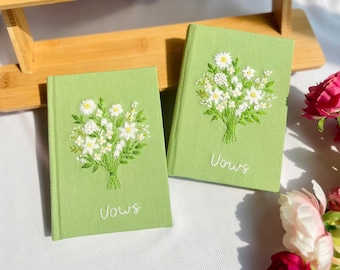 Puede incluir: Dos cuadernos verdes con flores blancas bordadas y la palabra "Vows" en escritura blanca. Los cuadernos están sobre una superficie de madera, con un ramo de flores rosas en el fondo.
