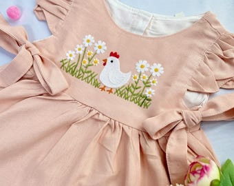May include: A light pink baby dress with a white embroidered chicken and daisy design. The dress has ruffled sleeves and tie details on the shoulders. The dress is made of a soft fabric.